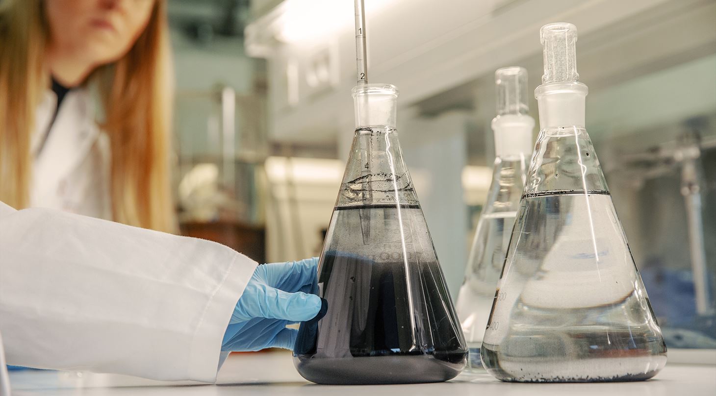 En laboratoriemedarbejder i hvid kittel og blå handske analyserer væsker i store koniske kolber. Den ene kolbe indeholder en mørk væske, de øvrige klart vand, hvilket illustrerer test og analyse af vandkvalitet.